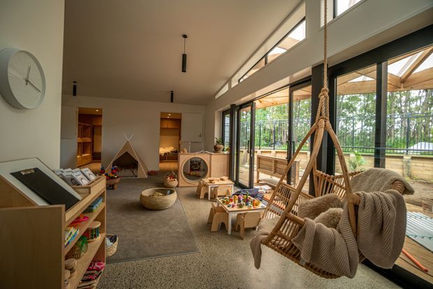 A bright, open-plan family living and play area with high ceilings and extensive floor-to-ceiling glazing. The room includes a hanging wicker chair with knitted throws, low children's tables and toys, a built-in timber shelving unit, a small reading nook, and a neutral carpeted seating area. Large sliding glass doors open to a timber deck and planted outdoor area, while clerestory windows bring in additional daylight. The interior has warm timber details, neutral finishes, and a relaxed contemporary feel.