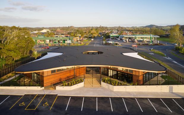 Aerial exterior view of a low-rise contemporary commercial building with a broad dark flat roof, warm timber-clad facade, extensive glazing, central entrance stairs, and landscaped planting beds. The building fronts a marked car park with accessible parking spaces and is situated within a suburban commercial setting.