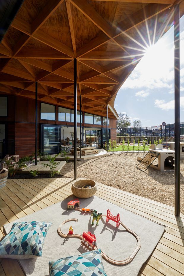 Sunlit covered outdoor play and lounge area beside a timber-clad building, featuring a broad exposed timber roof structure with triangular bracing, black slender posts, timber decking, large glazing, and a children's play setup on a light rug. A fenced landscaped yard and playground equipment are visible beyond.