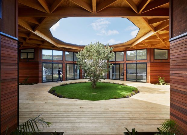 A circular or polygonal timber-lined courtyard with a central grass island and small tree, surrounded by a broad timber deck and glazed rooms. The architecture features rich dark timber wall cladding, extensive floor-to-ceiling glazing, exposed timber roof trusses, and a large octagonal roof opening that brings daylight into the courtyard. The image has a warm, natural, highly crafted character.