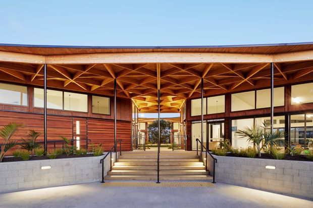 Front entrance courtyard of a low-rise contemporary building, featuring a broad timber canopy with an intricate exposed geometric timber ceiling, slender dark metal columns, extensive glazing, horizontal timber wall cladding, and raised concrete planter walls with tropical landscaping. A central stairway and accessible ramps lead to a glazed entry passage. The architecture is warm, refined, and strongly defined by its timber structure.