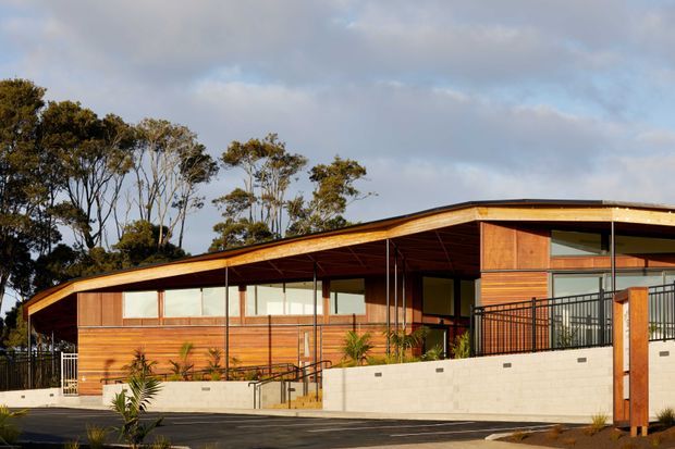 Single-storey contemporary timber-clad building viewed from the street, with a broad low-pitched roof, extensive glazing, covered entry and terrace areas, black metal balustrades, and a pale masonry retaining wall. Native trees surround the property, and the warm timber facade is lit by low-angle sunlight.