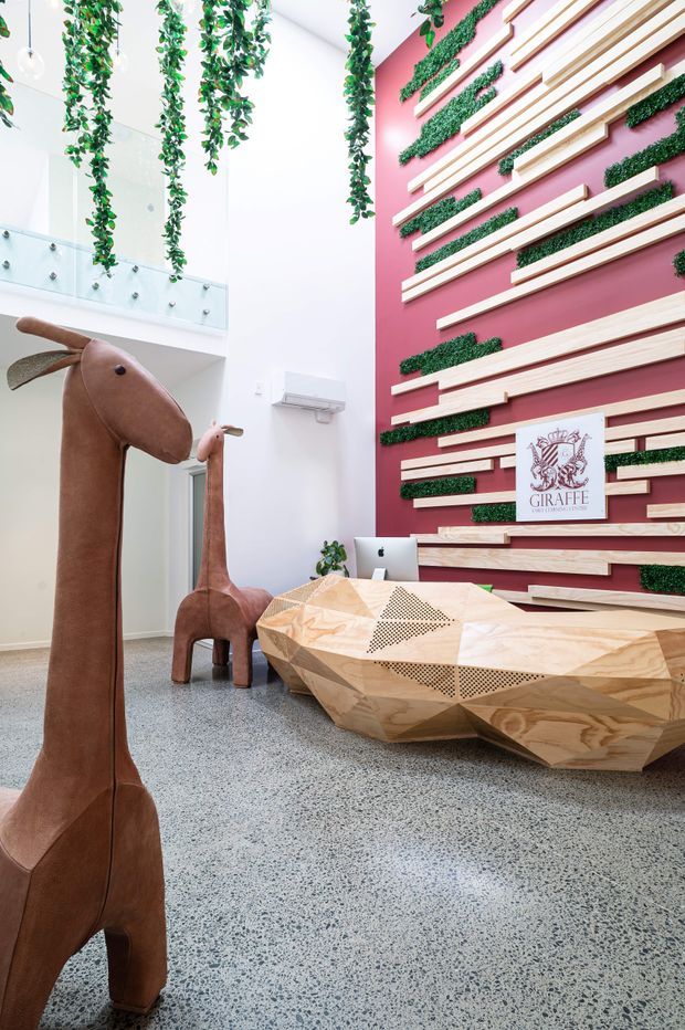 A bright, playful reception or commercial lobby with a high white ceiling and a glass balustrade overlooking an upper level. Hanging greenery descends from above, while a feature wall is painted deep pink and layered with horizontal light timber battens and strips of planted greenery. A sculptural faceted timber reception desk sits in front of the wall, with a computer and giraffe-themed signage. Two tall brown giraffe sculptures add a whimsical focal point. The floor is finished in speckled polished terrazzo.