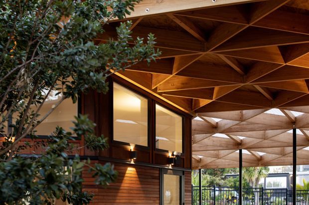 A covered outdoor architectural space with a dramatic exposed timber ceiling formed by repeating triangular and folded geometric beams. Dark timber-framed glazing and horizontal timber wall cladding define the adjoining building, while a mature tree partially screens the foreground and a sheltered terrace with metal balustrading is visible beyond. The scene has warm, sophisticated lighting and strong structural detailing.