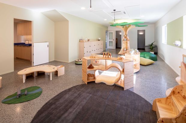 Bright, open-plan contemporary interior featuring a playful children's play and activity area. The room has polished terrazzo-like flooring, pale warm walls, a white ceiling with recessed lighting and air-conditioning, built-in cabinetry, a central timber-and-white island or activity table, sculptural timber play structures, low children's furniture, and a large dark circular rug. A compact kitchenette or utility area is visible through the opening on the left.