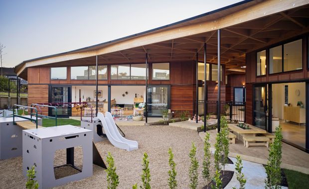 Exterior view of a contemporary low-rise building with a broad sloping roof and deep timber-lined eaves. Large glazed openings reveal an indoor communal or play area, while the foreground contains a landscaped gravel courtyard with a children's slide, play structure and steps leading into the building.