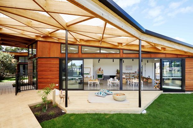 Single-storey contemporary pavilion-style home viewed from the garden, with extensive floor-to-ceiling glazing, a broad timber-framed pitched roof canopy, warm horizontal timber wall cladding, and open-plan dining and living areas visible inside. A timber deck and paved walkway connect the interior to the lawn and landscaped garden.
