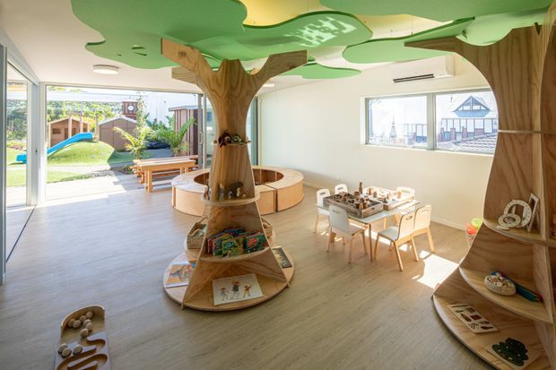 A bright, playful children’s learning and play space with a timber tree-themed interior. Sculptural tree trunks with integrated circular shelving and seating support books and toys, while green canopy-shaped ceiling panels create a whimsical forest atmosphere. The room includes a small activity table with child-sized chairs, pale timber-look flooring, large windows, and direct views to an outdoor playground.