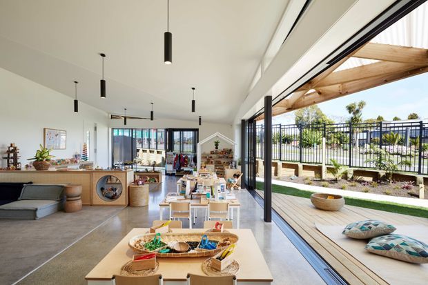 Bright, open-plan contemporary interior with a children's activity and play area, featuring extensive black-framed sliding glass doors opening to a timber-decked outdoor space. The room includes low tables and chairs, shelving, craft materials, a seating nook, polished concrete flooring, and a covered pergola beyond the glazing.