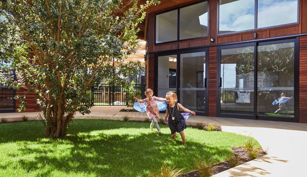 Exterior view of a contemporary multi-level home opening onto a small landscaped courtyard. The facade combines dark timber cladding and large dark-framed glazed doors and windows, with two children running across the lawn and a paved walkway in the foreground.
