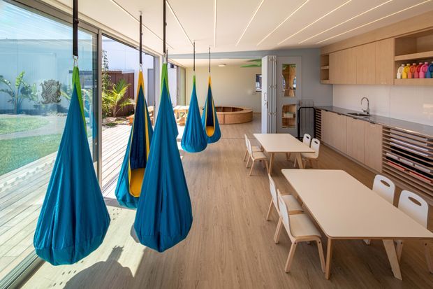 Bright contemporary multipurpose interior with full-height sliding glass doors opening to a landscaped courtyard. Several vivid blue and green hanging pod swings occupy the glazed edge, while light timber dining tables and white chairs sit alongside a long run of light timber cabinetry. The space includes a curved built-in seating nook and a calm, open-plan layout with abundant natural light.