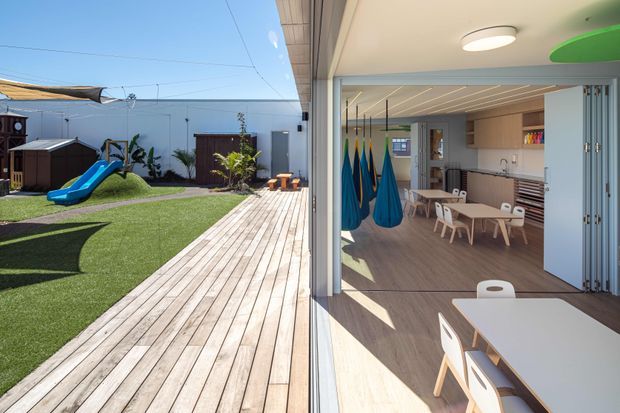 Bright view from a timber deck into an open-plan children's learning and activity space. The room has large sliding glass doors, timber-look flooring, pale wood cabinetry, a long kitchen or kitchenette bench, small tables and chairs, and colorful suspended pod swings. Outside is a landscaped play area with lawn, a slide, play equipment, and a broad timber deck.