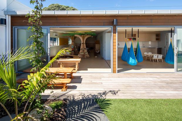 Bright view of a contemporary single-storey home opening onto a wide timber deck and landscaped garden. Large sliding glazed doors connect the deck to an open-plan interior with timber finishes, a dining area, and a children's play or recreation space featuring colourful hanging pod chairs. The flat roof, horizontal timber cladding, extensive glazing, and indoor-outdoor connection are prominent.