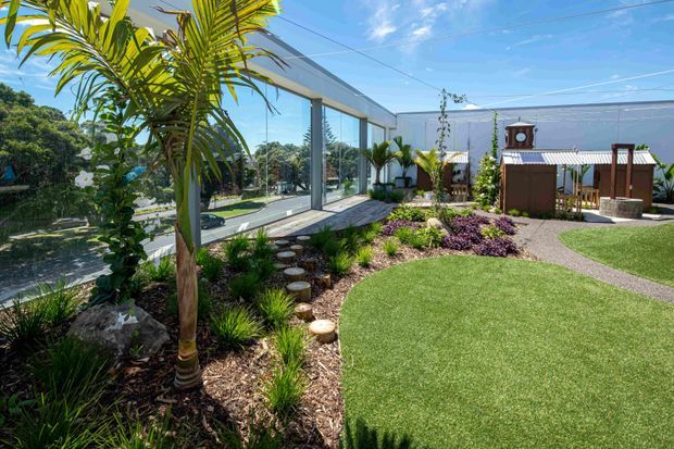 A landscaped outdoor courtyard or garden beside a contemporary building, featuring extensive floor-to-ceiling glazing, a covered outdoor sitting structure, curved artificial turf, planted garden beds, palm trees, stepping stones, and a bright blue sky. The setting has a relaxed subtropical feel with a clean, modern building edge and generous outdoor entertaining space.