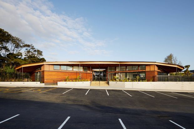A wide, low-slung contemporary building viewed from across a marked asphalt parking area. The facade combines warm horizontal timber cladding, extensive dark-framed glazing, a shallow projecting roof with deep eaves, and a central recessed glazed entrance. White rendered planter and retaining walls with sparse planting run along the front. Mature trees and blue sky form the background.