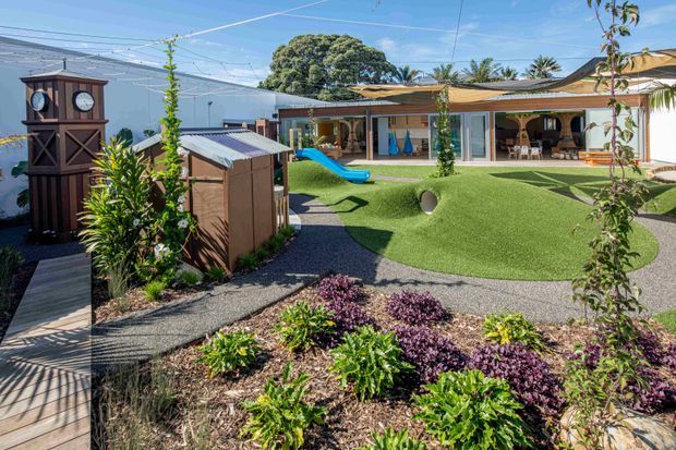 A bright landscaped courtyard or outdoor recreation area with artificial turf, curving pathways, planted garden beds, a children's slide, and a small timber playhouse or shed. A covered veranda with seating and glazed doors lines the single-storey building in the background. A tall decorative timber clock structure stands beside a boardwalk, with mature trees and overhead shade or utility structures visible around the site.