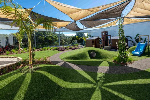 A sunny, landscaped outdoor play area with sculpted artificial grass mounds, winding dark pathways, a tunnel, blue slide, and timber play structures. Large suspended shade sails cover the space, with tropical planting and garden beds around the perimeter.