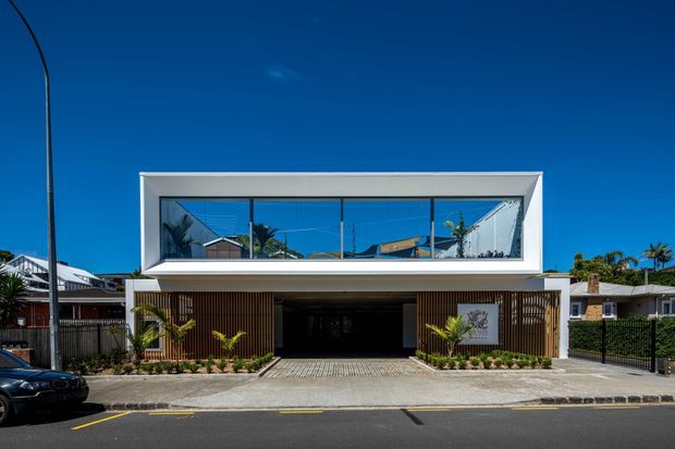 Street-facing contemporary two-storey building with a crisp white rectangular upper volume, expansive reflective glazing, and a recessed ground-level frontage screened by vertical timber battens. A wide paved driveway and dark garage or entry opening occupy the lower level, with neighbouring suburban homes and palm planting visible around it.