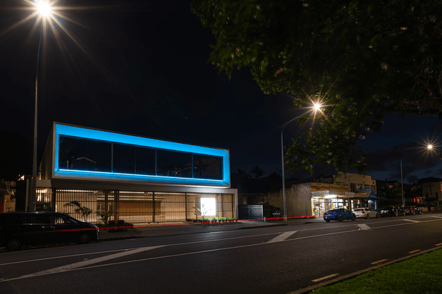 Night-time street view of a contemporary two-storey commercial building with a strongly illuminated blue-trimmed upper-level glazed box, a glazed and vertically screened ground floor, and bright interior lighting. The building fronts a wide urban road with streetlights, neighbouring shops, parked vehicles, and mature trees.