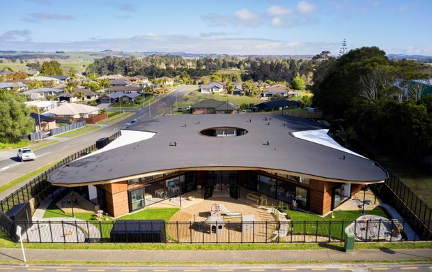 Aerial exterior view of a low-rise contemporary building with a broad curved flat roof, extensive glazing, timber-toned wall cladding and a fenced landscaped forecourt. The building sits in a suburban setting with residential streets and open countryside visible beyond.