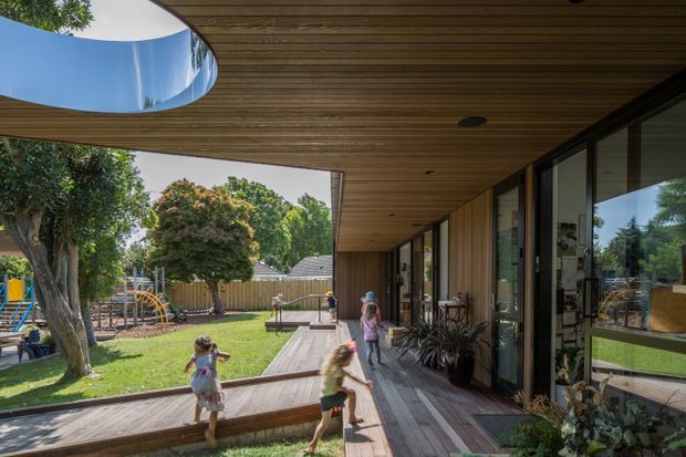 A covered outdoor deck or veranda runs along a glazed contemporary building, with a warm timber-lined ceiling, timber decking, full-height sliding glass doors, and a large curved roof opening framing the sky. The deck overlooks a landscaped garden and children's playground, with indoor spaces visible through the glazing. The architecture has strong indoor-outdoor integration and a clean, contemporary linear form.