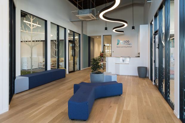 A bright contemporary reception area for an early learning centre, featuring pale timber flooring, white walls, extensive black-framed glazing, a white reception desk, blue curved modular seating, and distinctive sculptural black-and-white pendant lighting. Internal glazed rooms with decorative tree-patterned screening are visible along the left side.