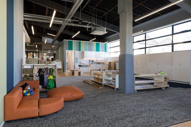 A spacious contemporary commercial showroom or retail interior with an open-plan layout, exposed services ceiling, linear pendant lighting, large industrial-style windows, and grey carpet. The space includes colourful striped wall cladding, white display shelving and counters, a clothing display area, and a bright orange modular lounge near the foreground. A structural concrete column divides the showroom, while a small kitchenette or service counter is visible toward the rear.