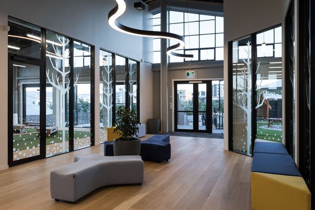 A bright contemporary commercial lobby or entry space with expansive black-framed floor-to-ceiling glazing, pale timber flooring, sculptural curved pendant lighting, and modern modular seating in grey, navy, and yellow. Decorative white tree-like graphics appear on the glazing, with an exterior courtyard and neighboring buildings visible beyond.