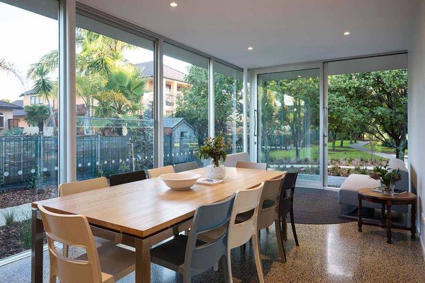 Bright glazed dining area with a large rectangular timber dining table and mixed modern chairs, surrounded by floor-to-ceiling windows and sliding glass doors overlooking a leafy garden. The room has a polished terrazzo-style floor, white ceiling with recessed downlights, and a small side table near the glazing.