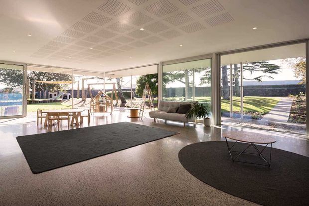 A bright, expansive contemporary living room or shared lounge with full-height corner glazing overlooking a landscaped garden and playground. The space has polished terrazzo or concrete-look flooring, a perforated acoustic ceiling grid with recessed downlights, neutral seating, dark area rugs, and a low geometric coffee table. The extensive glazing and open layout create a strong indoor–outdoor connection.