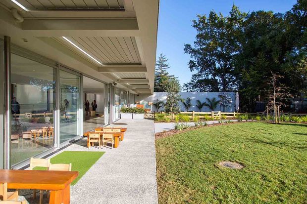 A bright contemporary building viewed from a covered outdoor terrace or walkway, with extensive floor-to-ceiling glazing opening onto a paved patio. Timber outdoor tables and benches sit beside artificial turf mats, while a landscaped lawn, planting beds, palms and a low rendered wall form the garden beyond.