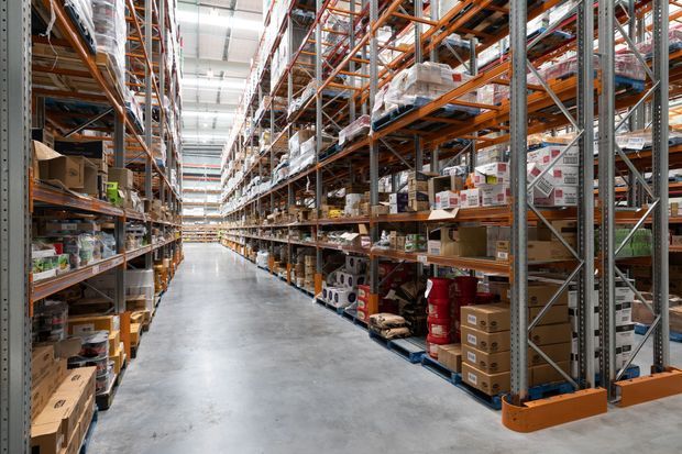 A large industrial warehouse aisle with tall orange-and-grey steel pallet racking on both sides, densely stocked with boxed goods, supplies, and pallets. The polished concrete floor and bright overhead linear lighting create a clean, functional commercial storage environment.