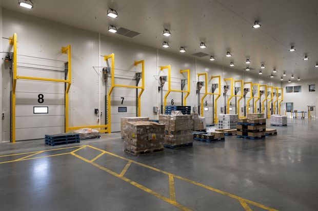 Large industrial warehouse or distribution facility interior with a polished grey concrete floor, high light-coloured insulated wall and ceiling panels, repeated loading-bay doors, bright yellow protective frames and floor markings, palletised goods, and rows of suspended industrial lights.