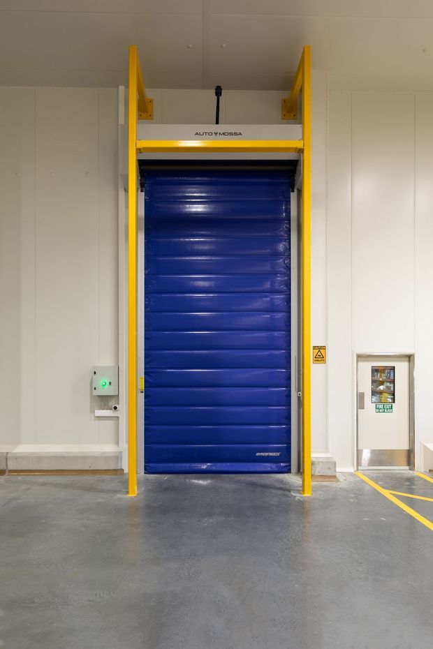 A commercial or industrial interior showing a blue high-speed roller door framed in bright yellow safety rails. The door is set into pale panelled walls, with a concrete floor, warning signage, a control panel, and marked safety lines leading to a small fire-exit door.
