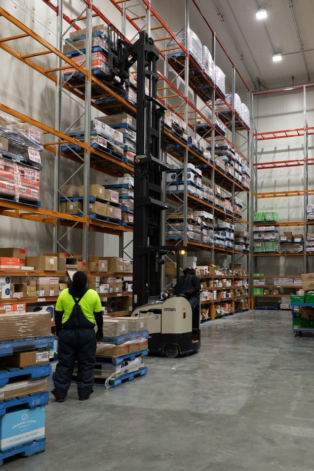A high-bay warehouse interior with tall orange-and-grey pallet racking fully stocked with boxed and palletised goods. A Crown forklift is operating in the central aisle, with two warehouse workers nearby. The space has a polished concrete floor, pale industrial walls, exposed overhead services, and bright suspended lighting.