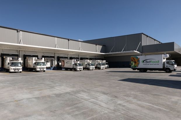 Exterior view of a large commercial food distribution warehouse with corrugated metal-clad building volumes, a broad loading dock canopy, multiple delivery trucks, and an expansive concrete service yard.