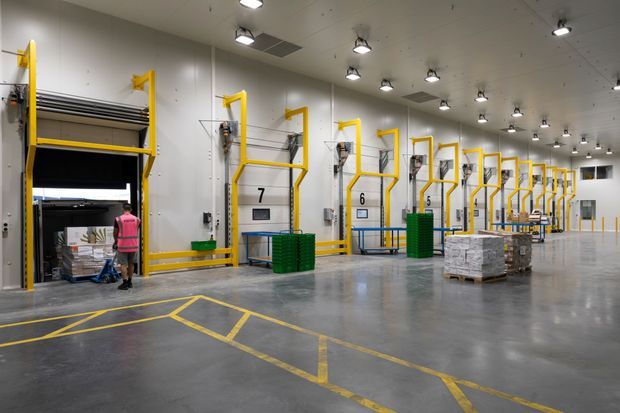 A large, brightly lit industrial warehouse or logistics facility interior with a long row of loading bay doors and yellow safety frames. The polished grey concrete floor is marked with yellow traffic and loading lines, while pallets, stacked goods, green barriers, and a worker in safety clothing reinforce the distribution-centre setting.