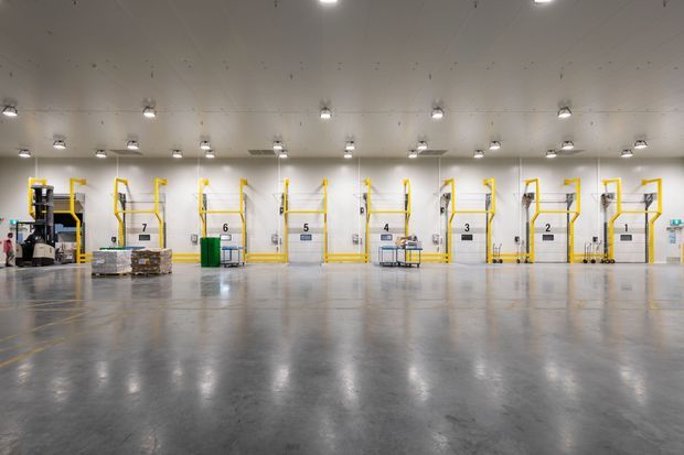 Wide interior view of a large industrial warehouse or logistics facility with polished concrete flooring, white walls and ceiling, extensive overhead lighting, and multiple numbered loading or storage bays with bright yellow safety rails and access platforms. Pallets, equipment, and industrial machinery are arranged along the perimeter.