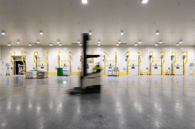 Wide-angle view of a large industrial warehouse or logistics facility with numbered loading bays, yellow safety railings, palletised goods, bright overhead lighting, and a forklift moving across a polished concrete floor.