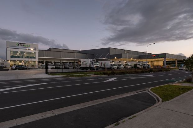 Exterior twilight view of a large contemporary commercial and industrial facility, with a dark metal-clad warehouse or distribution building, glazed office entrance, loading and delivery vehicles, landscaped frontage, and a marked roadway in the foreground.