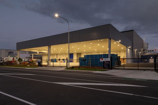 Exterior dusk photograph of a large contemporary commercial or industrial building. The structure has a broad flat-roofed canopy, dark horizontal ribbed cladding, extensive open-front glazing, bright recessed ceiling lights, loading or service areas, security fencing, and a marked urban roadway in the foreground.