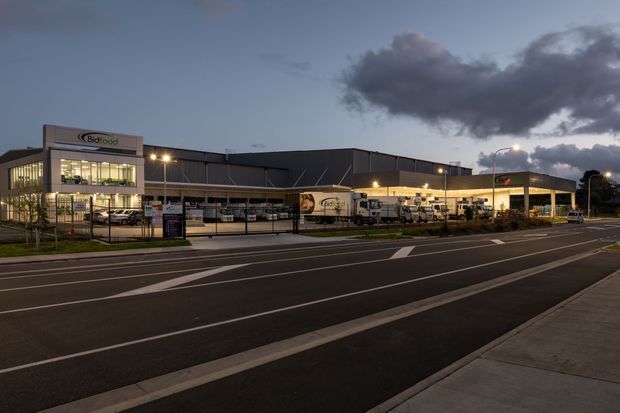 Wide dusk exterior view of a large commercial food-processing or distribution facility, with illuminated office glazing, dark metal-clad warehouse volumes, loading bays, trucks, signage, perimeter fencing, and a marked multi-lane roadway in the foreground.
