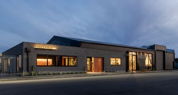 A dusk exterior photograph of a low-rise contemporary commercial building with a long, dark brick facade. Large illuminated windows and glazed entry doors reveal warm interior lighting, while vertical metal fencing and restrained landscaping line the street frontage. The building has a flat/low-pitched roof composition and a horizontal, single-storey form.