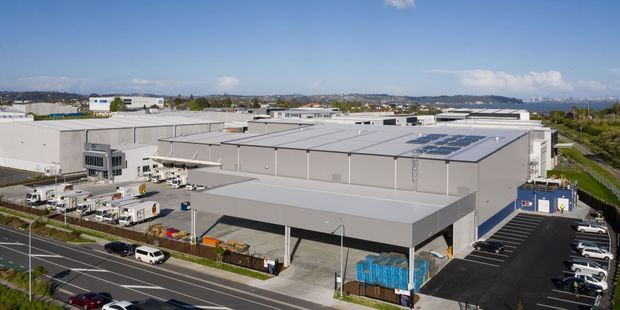 Aerial view of a large modern industrial or commercial warehouse complex with expansive light-grey metal-clad buildings, flat roofs, loading docks, service yards, marked access roads, parking areas, and rooftop solar panels. The surrounding suburban industrial landscape and distant water or hills are visible in the background.