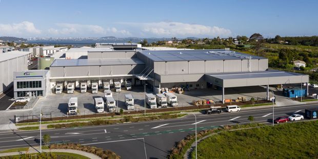 Aerial view of a large modern industrial or commercial warehouse and distribution facility, with extensive grey metal-clad buildings, loading docks, delivery trucks, paved service yards, parking, and a prominent multi-lane road in the foreground. The surrounding landscape includes low-density development, vegetation, distant hills, and a bright blue sky.