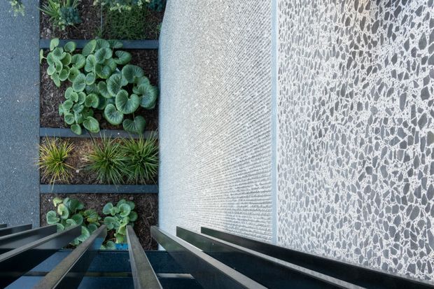A contemporary architectural detail viewed from above, showing a narrow landscaped courtyard or garden beside a tall textured wall. Dense low planting is arranged in rectangular garden beds separated by dark linear edging, while a patterned pebble-like feature wall and a finely textured pale wall form the architectural enclosure. Dark metal or timber structural fins project into the foreground, creating strong perspective lines.