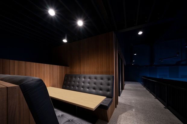 A dark, sophisticated cinema or theatre lounge with black upholstered booth seating, a light timber table, and warm vertical timber wall panelling. The space opens onto a carpeted circulation walkway with black railings and deep blue-lit areas beyond. Recessed and surface-mounted ceiling lights create a moody atmosphere.