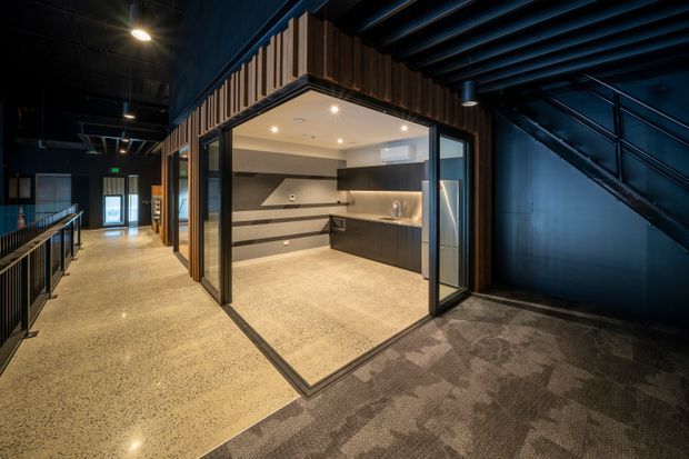 A modern commercial interior showing a compact kitchenette or refreshment area enclosed by black-framed glass walls and doors. The space features dark cabinetry, a stone-look benchtop, a linear tiled or panelled splashback, warm timber slat cladding above the enclosure, polished concrete flooring, and a dark exposed ceiling with a nearby steel stair. The surrounding circulation area has carpet and metal balustrading.