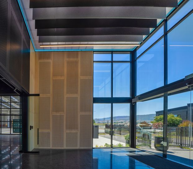 A dramatic double-height contemporary interior entry or lobby with extensive floor-to-ceiling glazing, dark exposed ceiling baffles, black-framed glass, and a large perforated timber feature wall. Blue accent lighting outlines the ceiling, while the polished dark floor reflects the daylight and views toward landscaped grounds and distant hills.