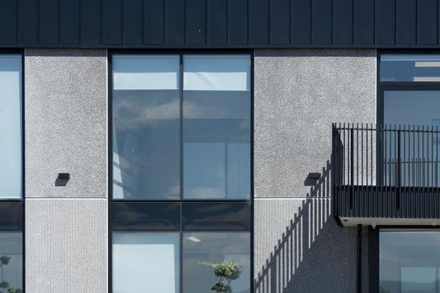 Close-up exterior view of a contemporary multi-storey facade with dark vertical metal cladding, textured light-grey wall panels, tall blue-grey glazed windows, and a black metal balcony balustrade. Strong geometric lines and shadows define the composition.